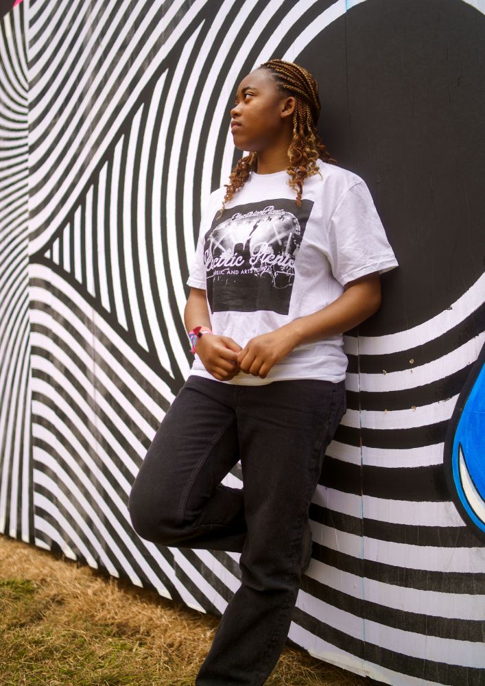 A person with braided hair stands against a black-and-white striped mural, wearing the Electric Picnic White Stage T-Shirt by Electric Picnic and black jeans, looking to the side thoughtfully.