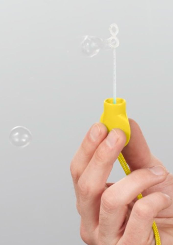 A hand holds the Electric Picnic Bubbles wand by Electric Picnic, creating translucent bubbles with its soapy loop against a plain, light gray background.